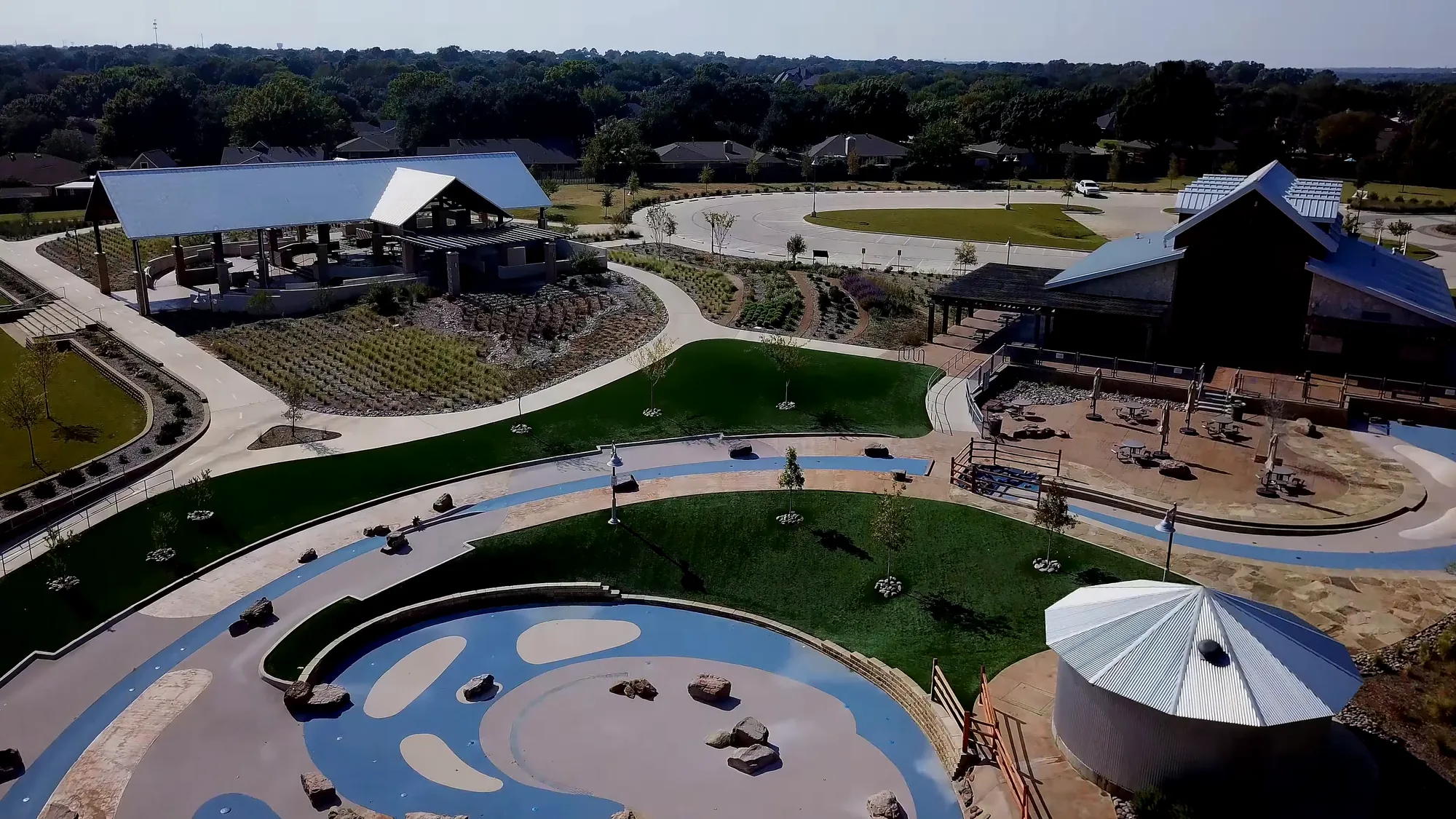Drone view over a finished park: a round blue and tan splash pad with boulders in front, a round metal roofed structure at right, a barn and an open pavilion behind green lawns and planted beds, with a parking loop and neighborhood trees beyond.