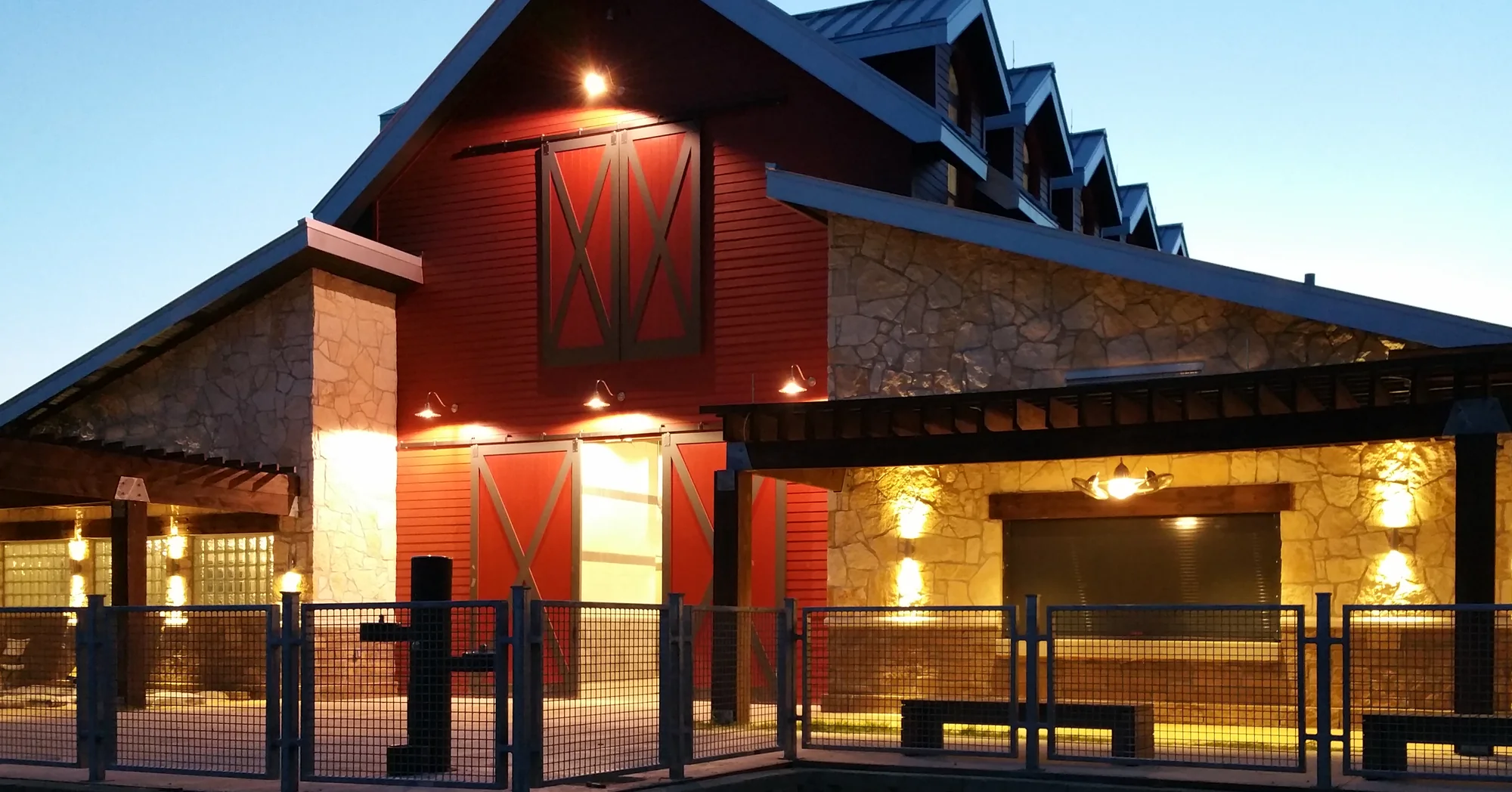 A red barn gable with a sliding hay door and lit barn doors below, stone walls and timber pergolas on both sides, a row of dormers along the roof and warm wall lights against a deep blue evening sky, behind a metal mesh railing.
