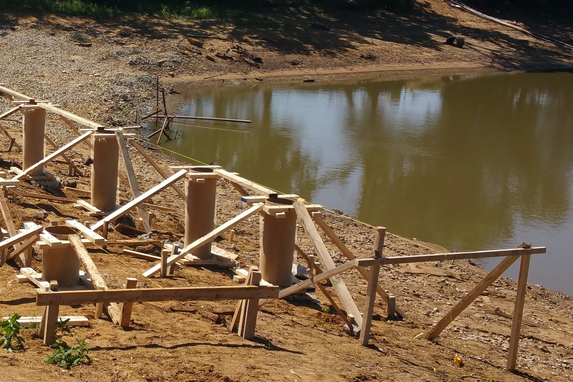 Cardboard tube pier forms braced with lumber and string lines on a sloped dirt bank, stepping down toward a brown pond with a gravel shore and trees reflected in the water.