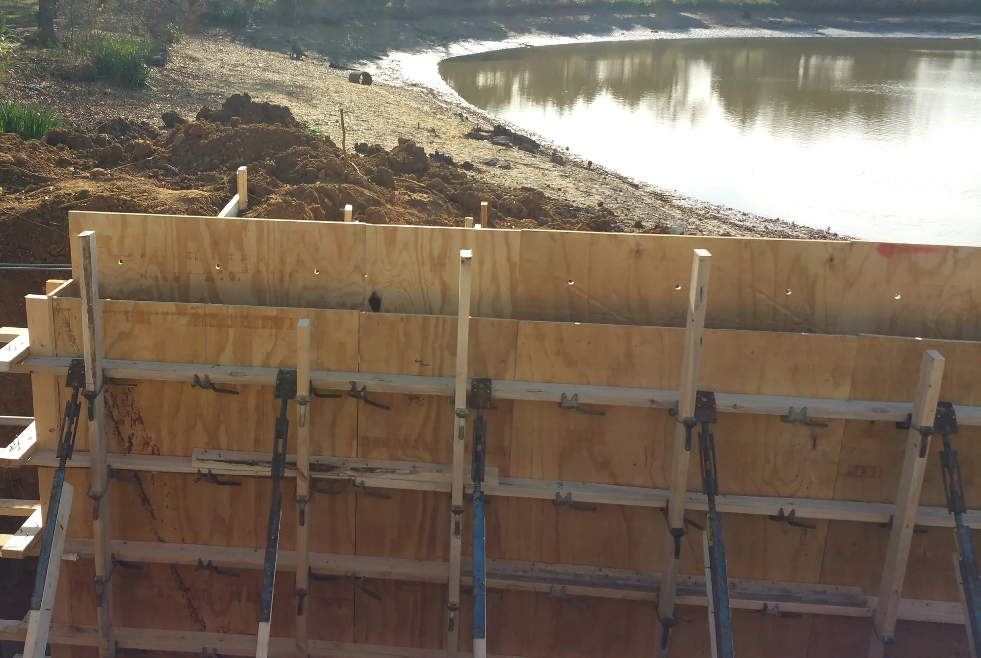 A long plywood wall form in an excavation beside a pond, held by horizontal wood walers, rows of metal form tie brackets and blue steel pipe braces, with dug-out soil and a muddy pond bank behind it.