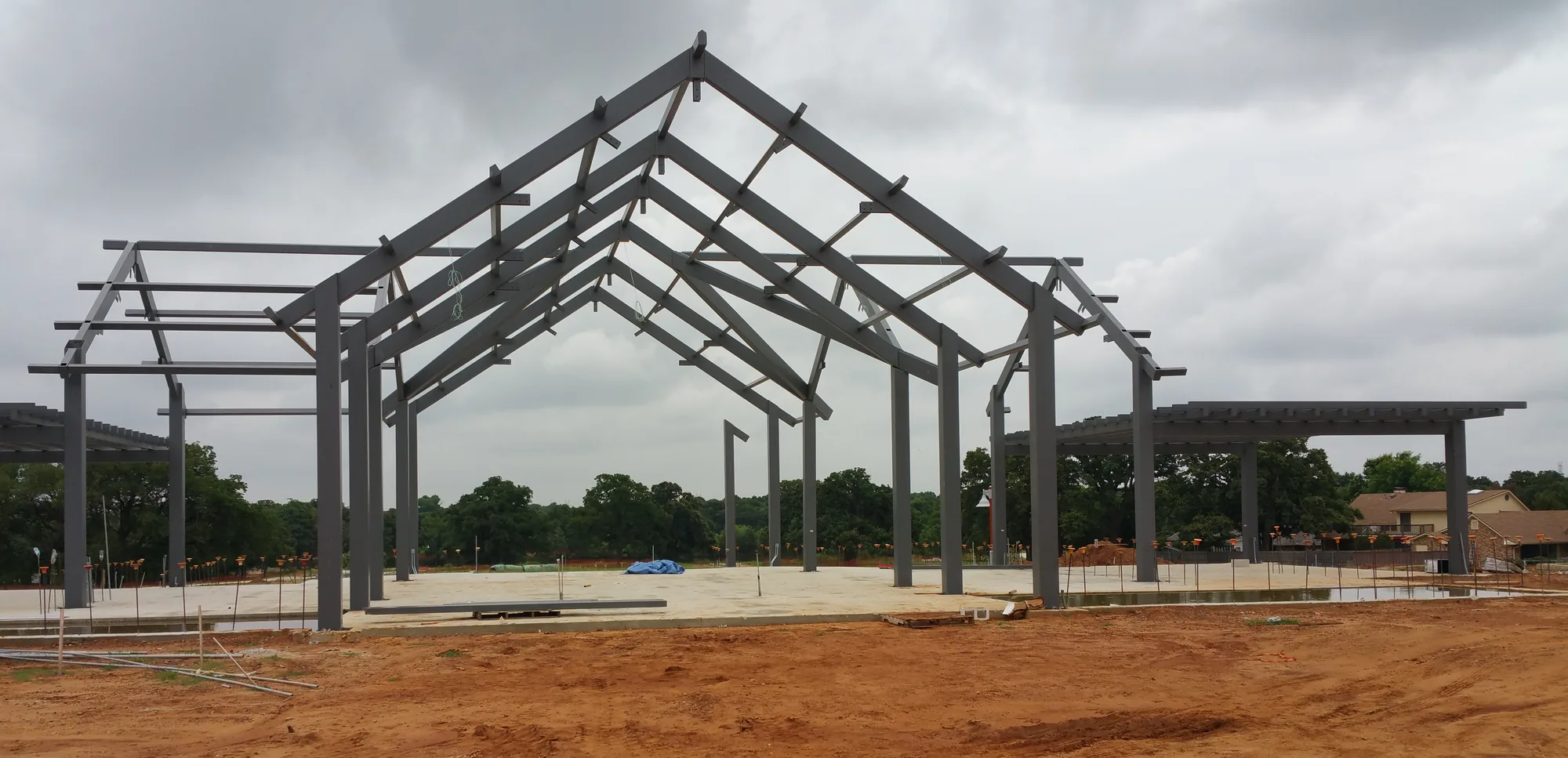 Gray steel gable frames and side pergola beams of an open pavilion rise over a concrete slab under a cloudy sky, with red dirt in the foreground and trees and houses behind.