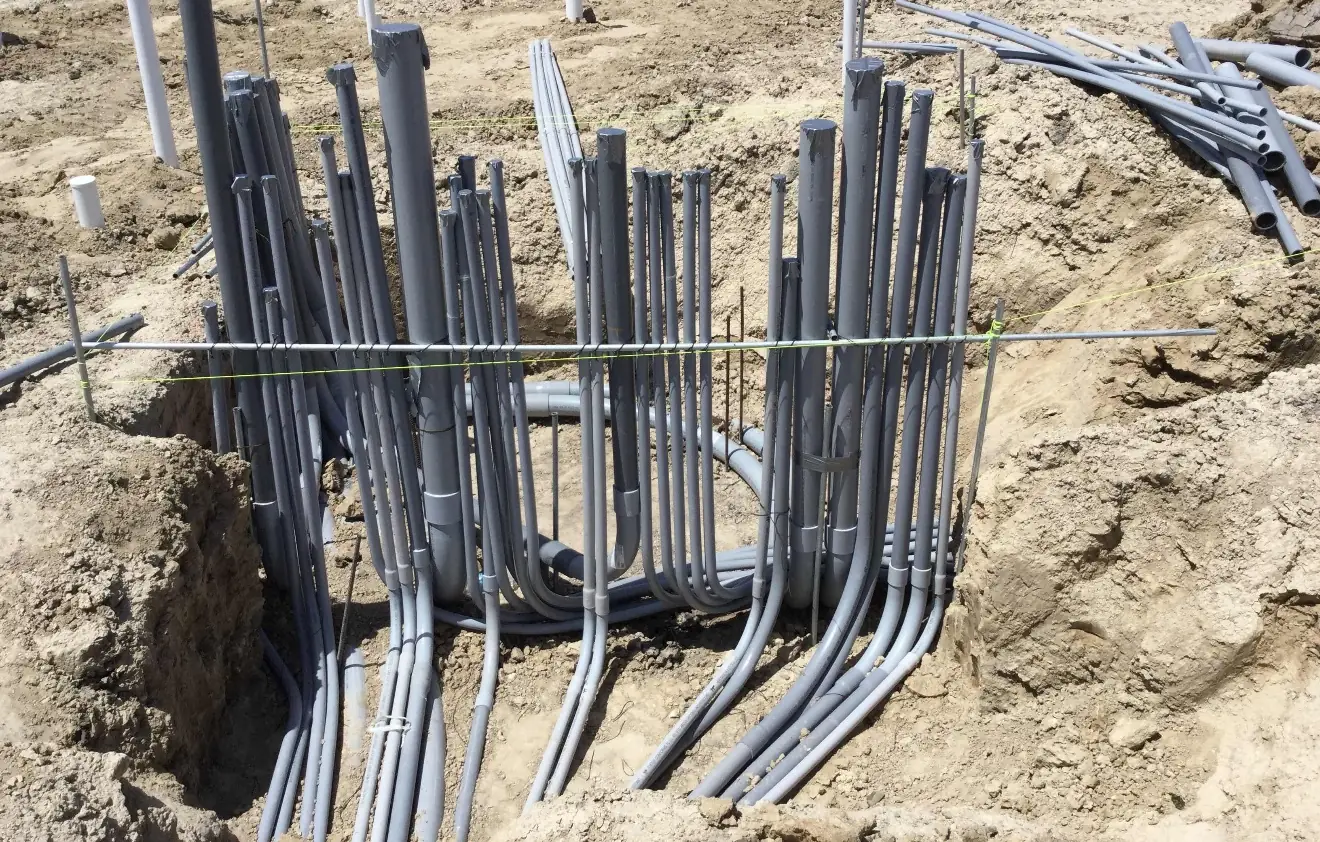 Two curved bundles of grey PVC conduit rise from a sandy excavation and are tied to a horizontal steel bar, with more conduit lengths piled at the upper right and white plumbing stub-ups in the dirt behind.