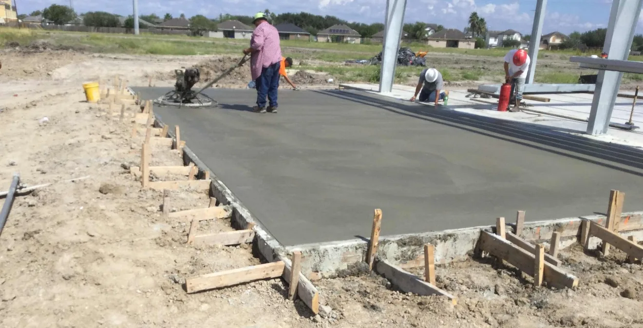 A crew finishes a fresh concrete pad edged with wood forms: one worker runs a power trowel, two kneel at the far edge beside galvanized steel columns, and houses line the field behind.