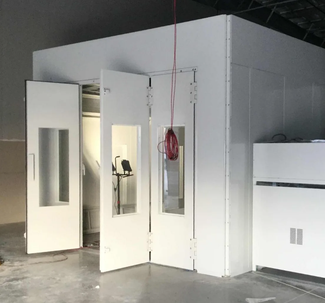 A large white paint booth with its tall doors swung open stands on a bare concrete floor inside an unfinished metal building, with a coiled red extension cord hanging in front, a white equipment cabinet at the right and a wrapped pallet at the lower left.