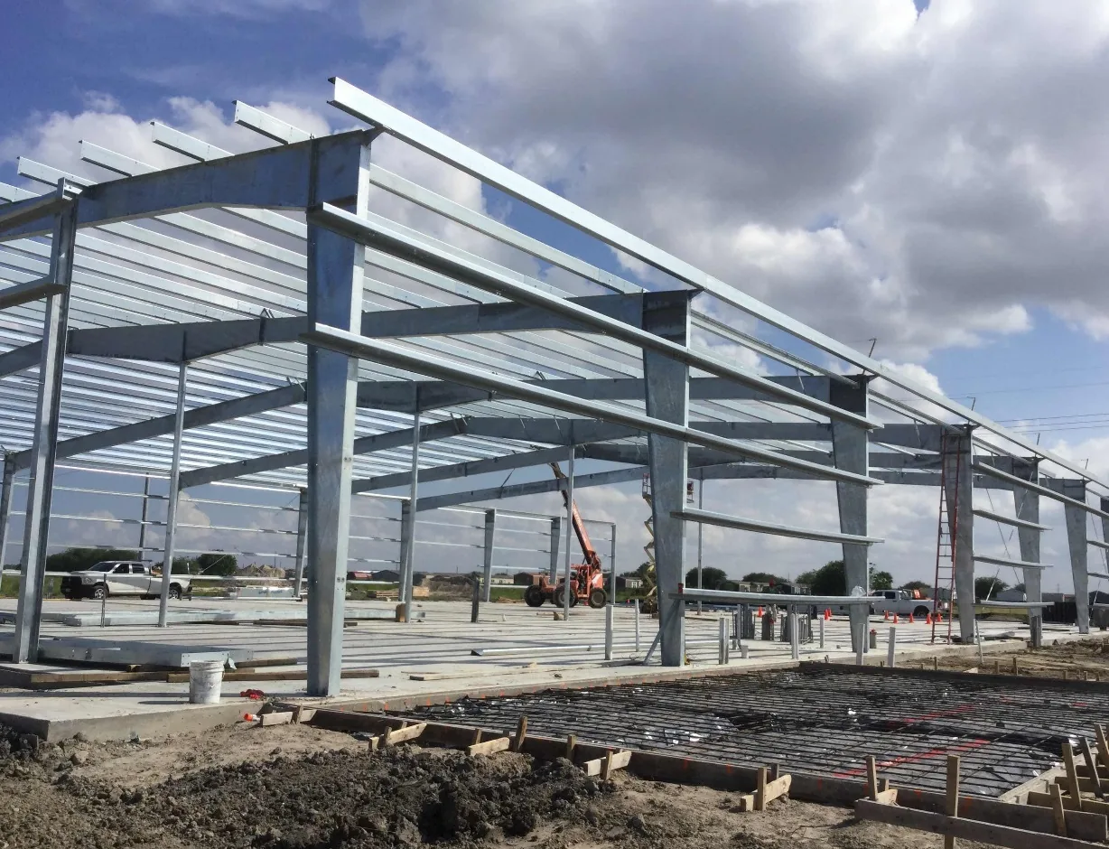 Galvanized steel columns, rafters and purlins of a pre-engineered metal building stand on a finished slab under a cloudy blue sky, with a rebar mat over black vapor barrier and wood forms in the foreground and a telehandler and scissor lift working inside the frame.