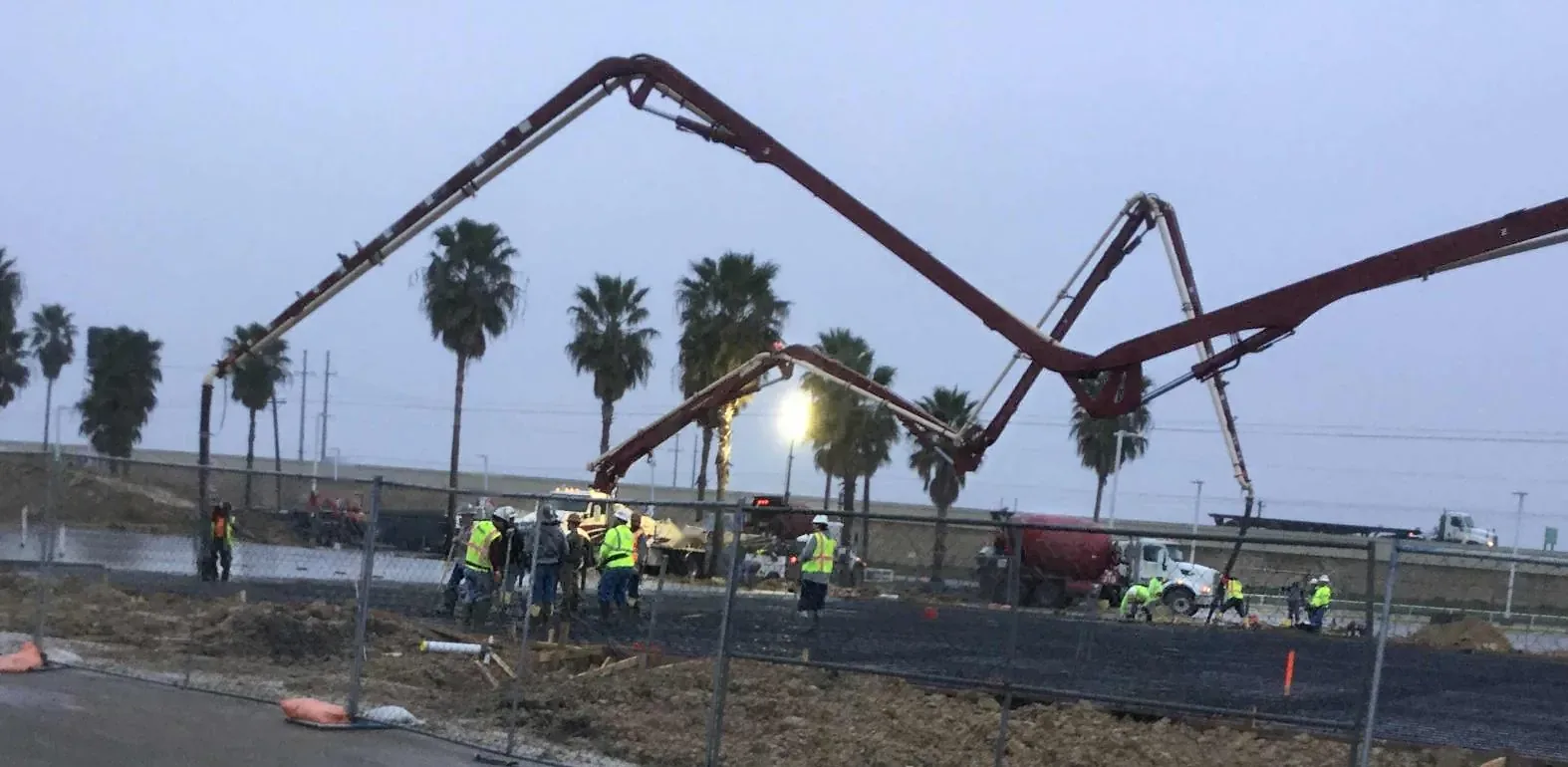 Red concrete boom pumps arch over a rebar mat at dusk while a crew in safety vests places concrete behind a temporary fence, with palm trees and a highway behind.