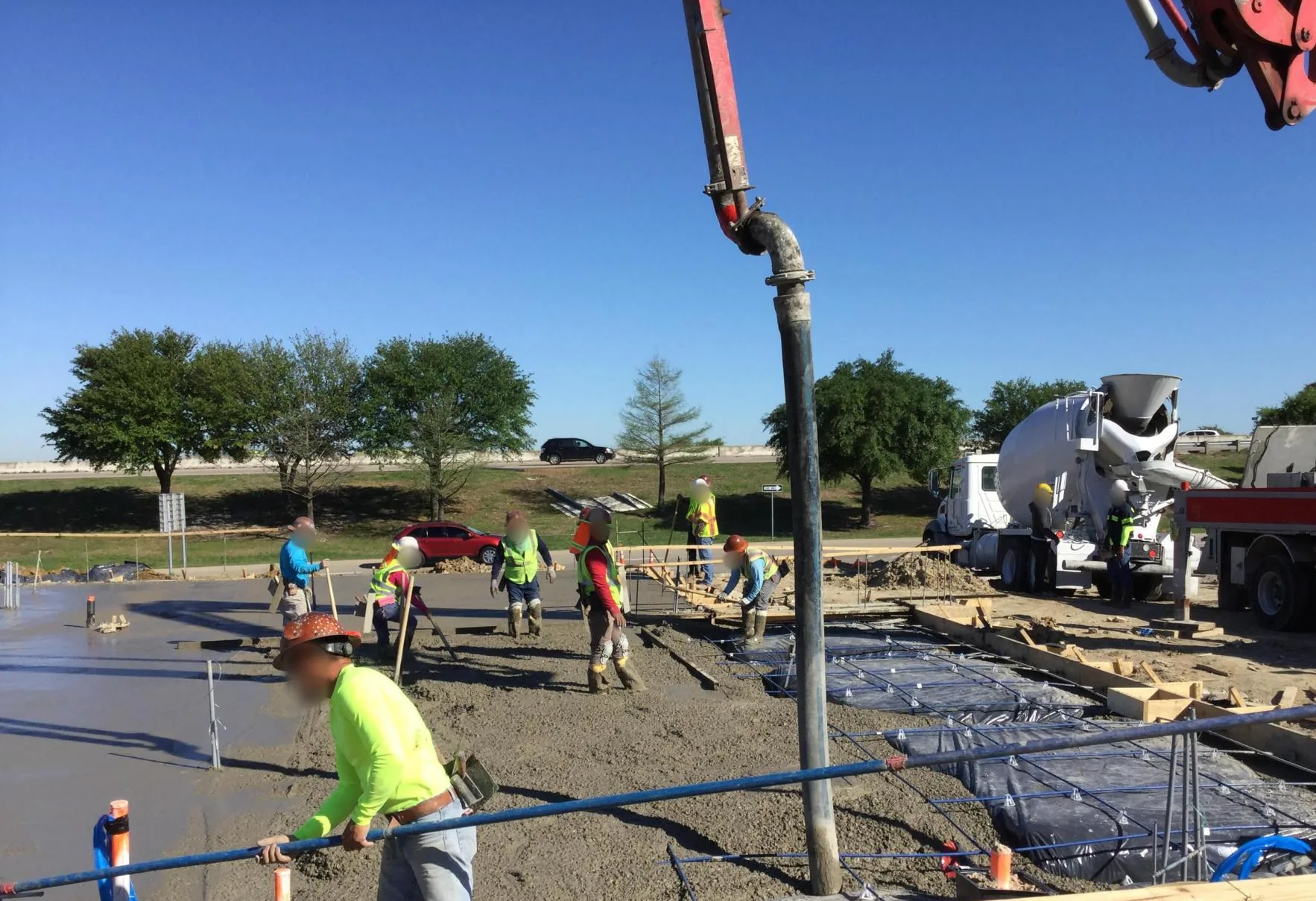 Under a clear blue sky, a concrete pump hose hangs over a crew in safety vests spreading fresh concrete across a slab, with blue reinforcing on chairs over black vapor barrier in the foreground and a mixer truck and pump truck behind.