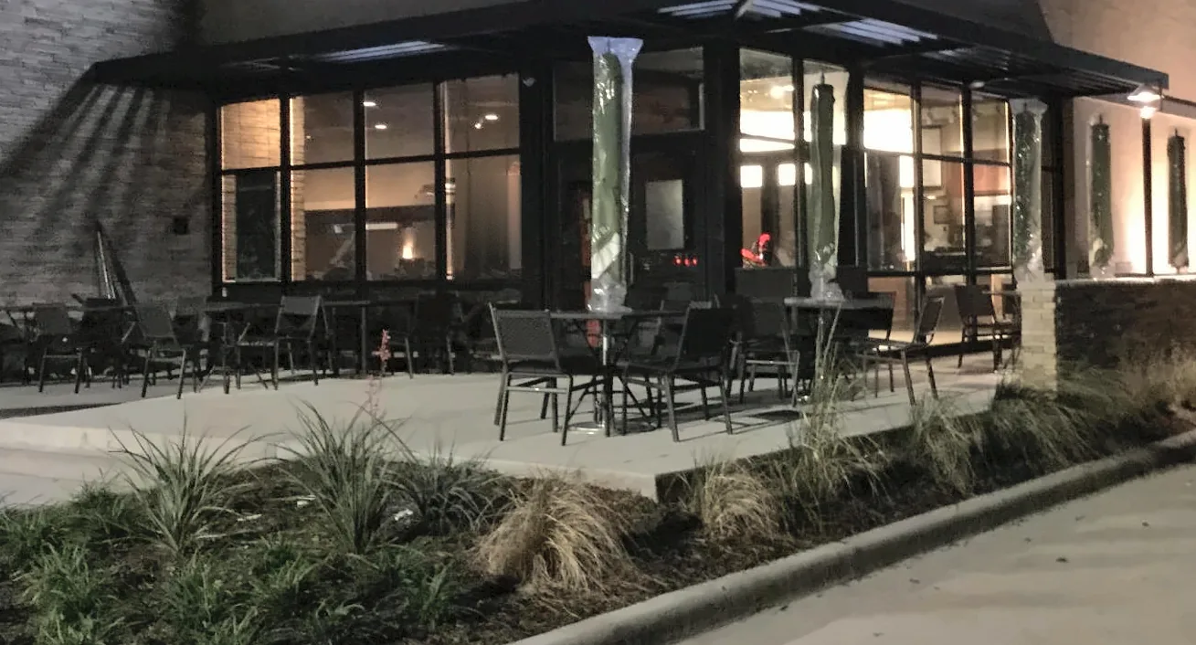 A restaurant at night: lit storefront windows under a dark steel canopy, a stacked stone wall at the left, patio tables and chairs with wrapped umbrellas, and new grasses in the planting bed along a curving curb.