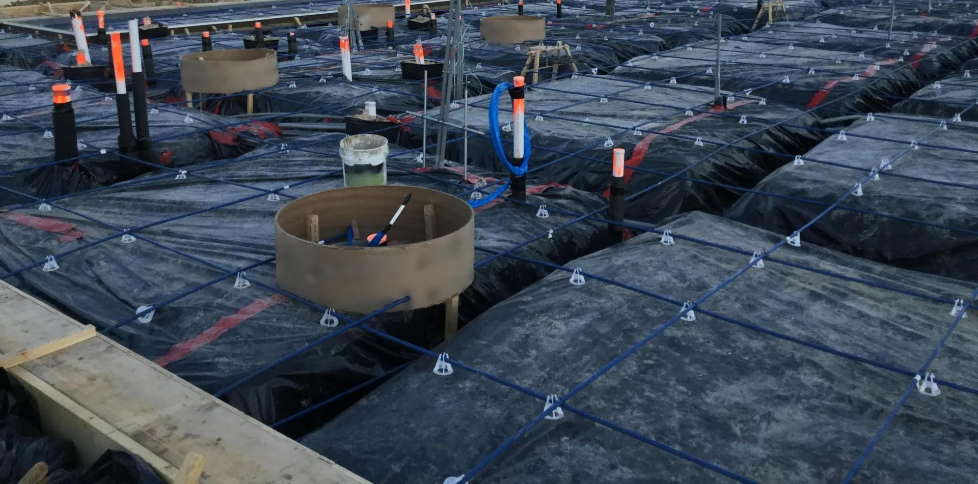 Blue post-tension tendons on small white chairs run in a grid over black vapor barrier and the trenches between pads, with round cardboard forms, orange-capped pipe stub-ups, a white bucket, a loop of blue tubing and a timber form edge at the lower left.