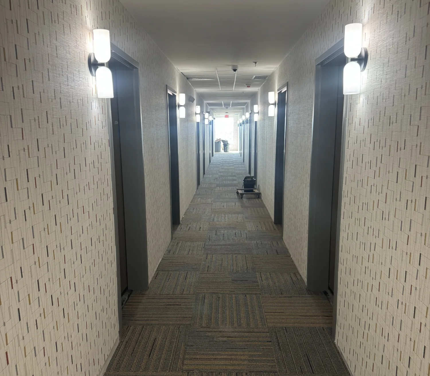A long, symmetrical hotel corridor with patterned wallcovering, paired white wall sconces beside guest room doors, and striped carpet tile running to a bright window at the far end.