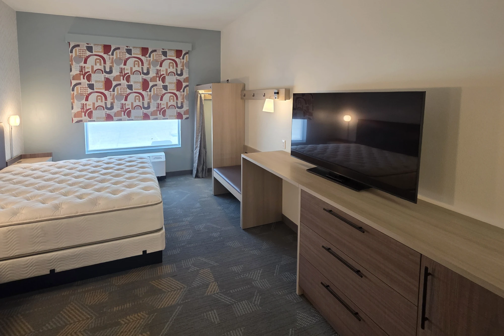 A hotel king studio seen from the entry side: a bare king mattress at left under a wall lamp, a window with a red, orange and navy patterned roller shade on a blue-gray accent wall, a wall unit below the window, an open wood closet with a gray curtain, a bench, a long light wood desk and dresser with a large black TV along the right wall, and gray patterned carpet.