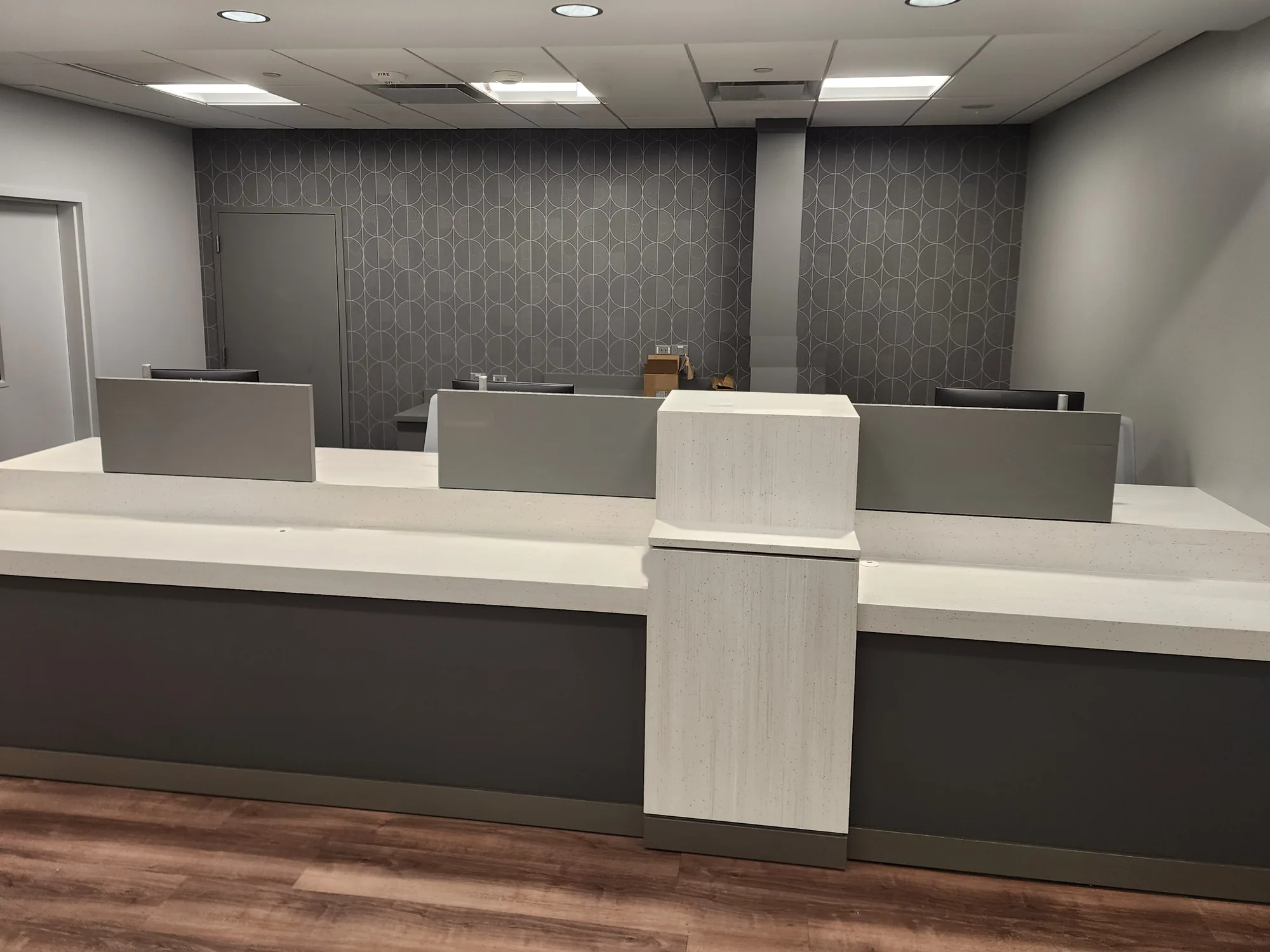 After: Teller counter with white quartz tops, gray privacy panels, and a light wood pedestal in front of a gray patterned wallcovering, under recessed lights.