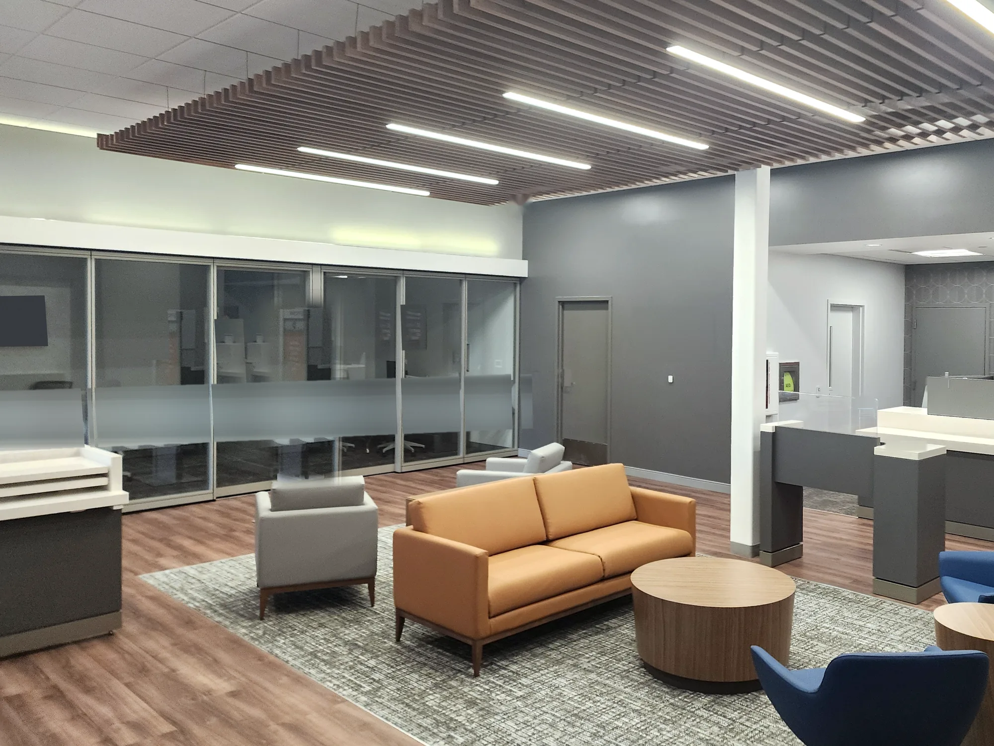 Daylit bank lobby with an orange sofa, gray and blue lounge chairs, and round wood tables on a patterned rug, glass-front offices at the left, a teller counter at the right, and a wood slat ceiling with linear lights.