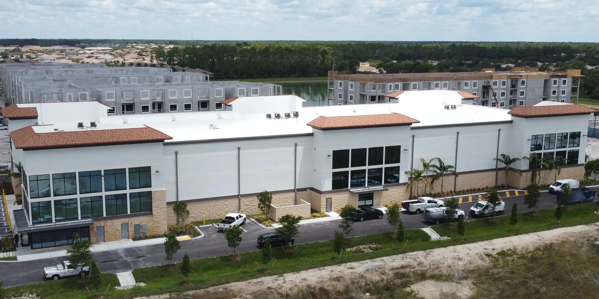 Low aerial of a long white three-story storage building with terracotta tile roof accents, glass corner towers and a stone base, palms and service trucks along the drive, apartments under construction behind and a sandy lot in front, under a cloudy sky.