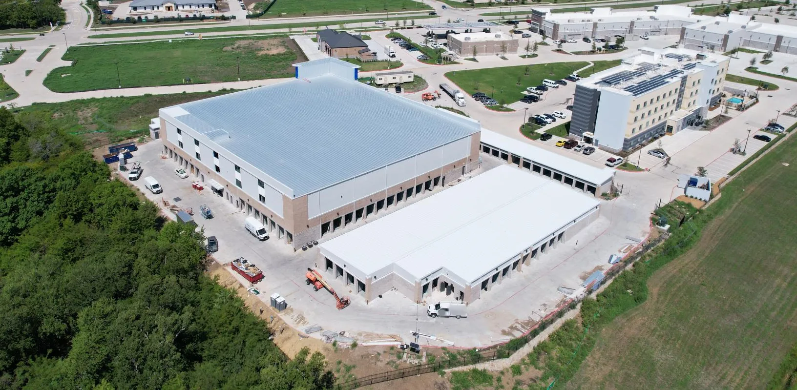 Aerial view of a large three-story storage building with a light metal roof and brick base beside long single-story drive-up buildings, a boom lift and service trucks in the drive aisles, woods at left and a hotel and shopping center behind.