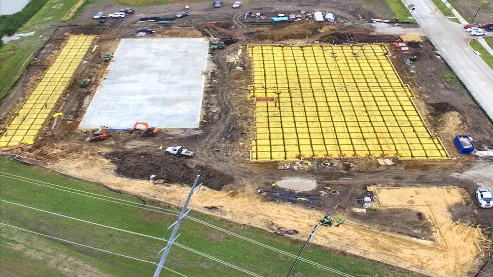 Aerial view of the site with one gray concrete building slab poured between two large yellow grids of foundation prep, an orange excavator, a skid steer, pickups and a blue dumpster around them, the pond at upper left, the new street at upper right and a power line across the grass in front.