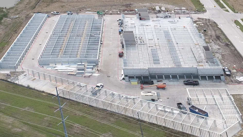 Aerial view of the same site with silver metal framing standing on the slabs: long single-story buildings at left and across the front and the main building at right with concrete stair and elevator shafts, an orange boom lift, a white storage container, a dumpster and pickups in the drive aisles, the pond at upper left, the new street at upper right and the power line across the front.