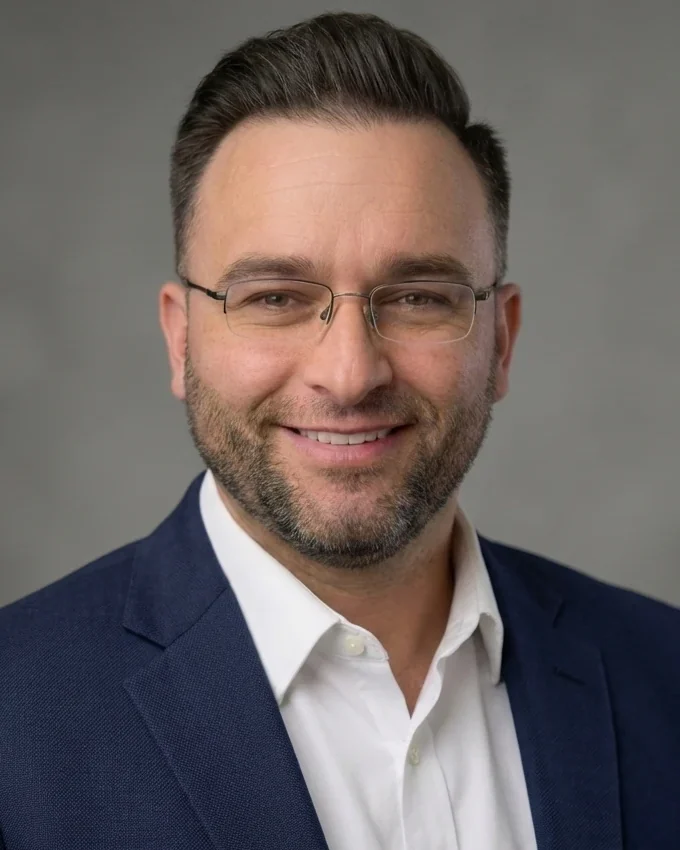 Head-and-shoulders photo of Austin Jones smiling, wearing glasses, a navy blazer and a white open-collar shirt, against a gray studio backdrop.