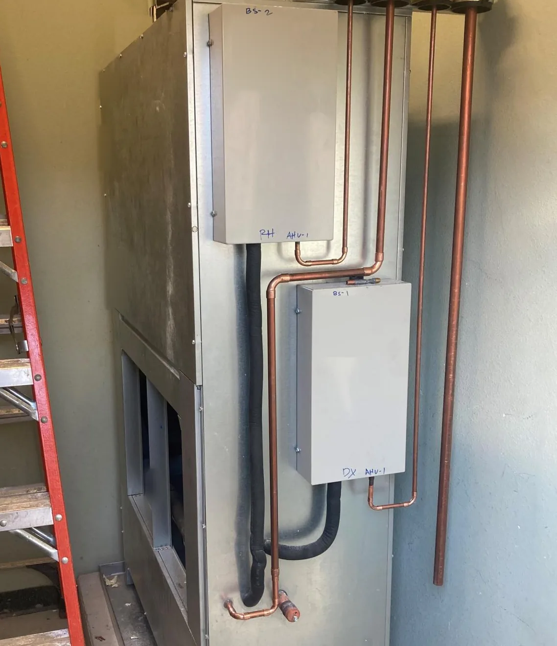 Tall galvanized air handling cabinet in a narrow mechanical room with two gray branch selector boxes on its front, marked BS-2 RH AHU-1 and BS-1 DX AHU-1 in marker, copper refrigerant piping and black insulated lines running up out of the frame, and an orange ladder at the left edge.