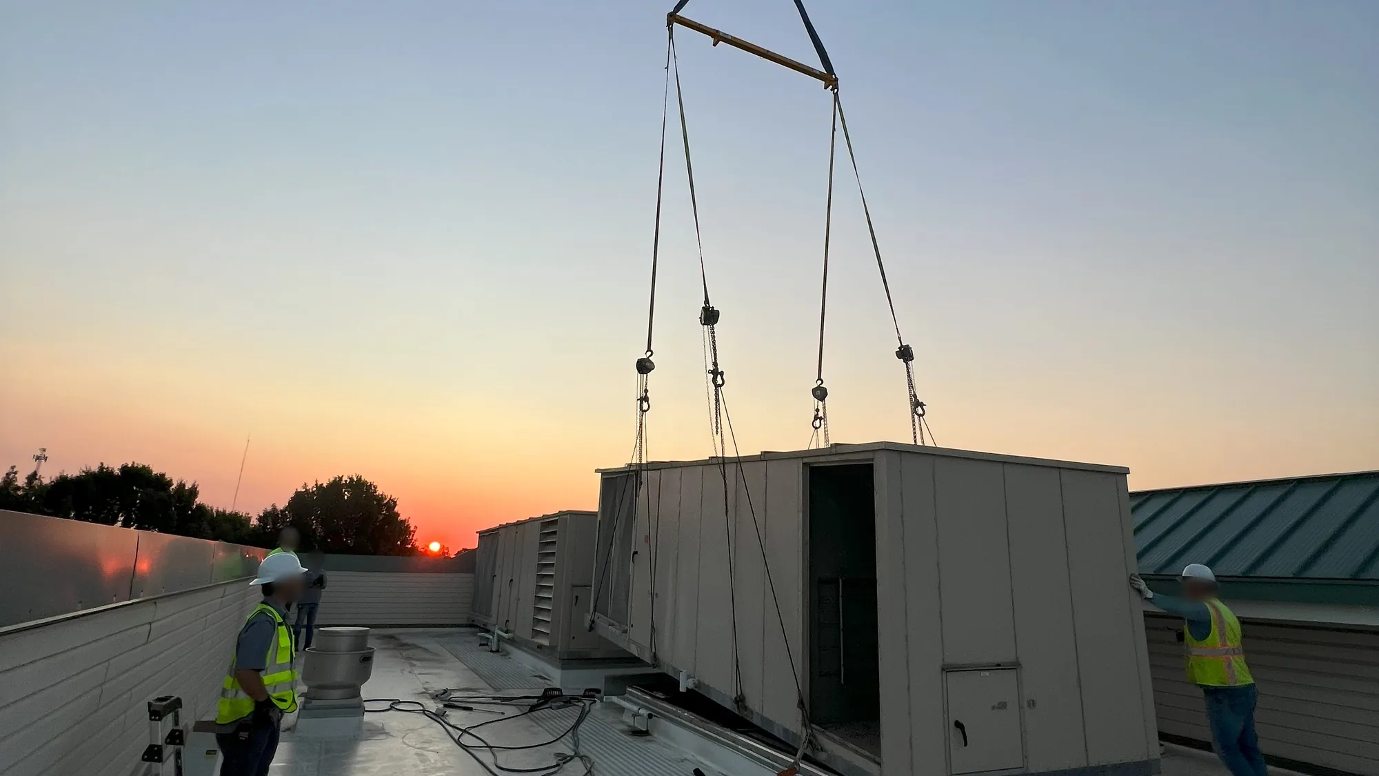 Sunset over a flat white roof where a crane holds a tan rooftop unit on four straps and a spreader bar, a crew member on the right steadies it by hand and three people stand on the left.