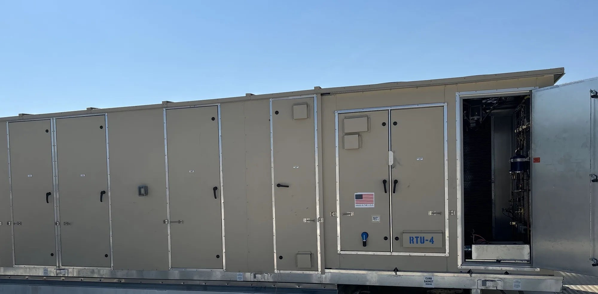 Long tan rooftop unit labeled RTU-4 on its roof curb under a clear blue sky, running off the left edge of the frame, with a row of access doors, a flag sticker on the control doors and an open galvanized door on the right showing refrigerant piping inside.