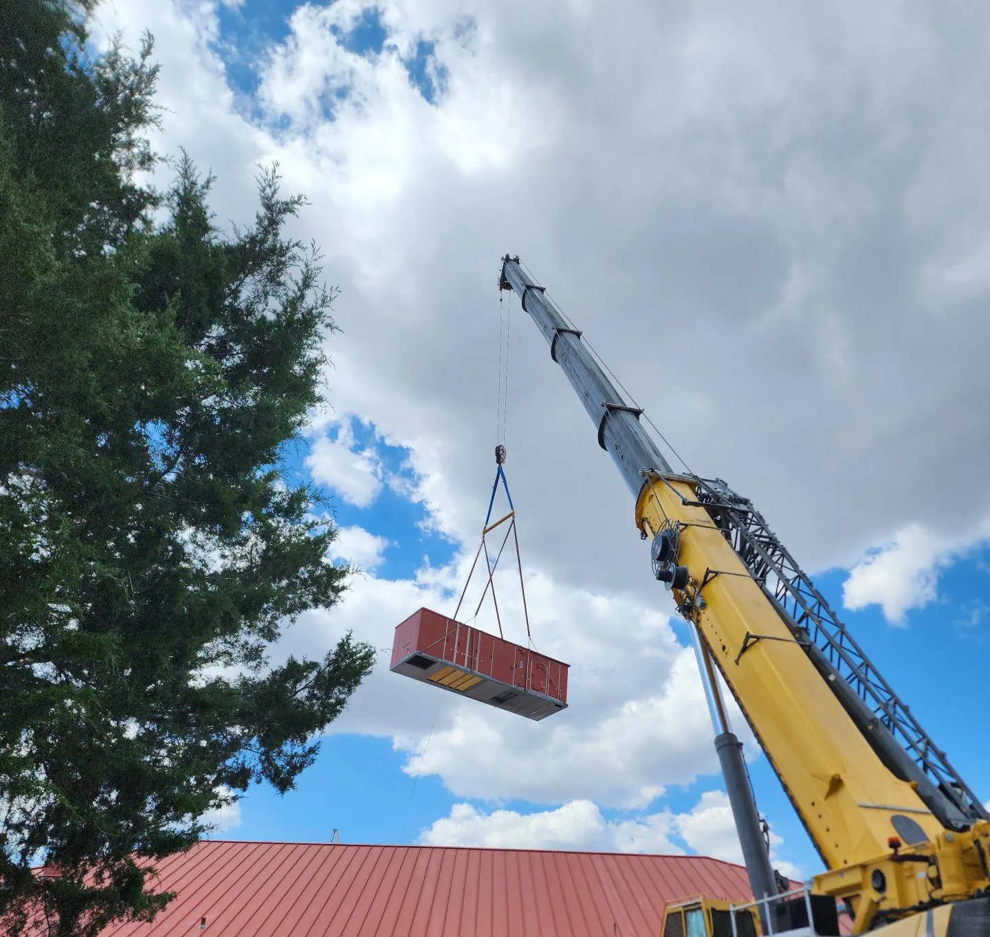 Yellow mobile crane lifting a rooftop unit high above a red standing seam metal roof, with a tall evergreen tree on the left and a partly cloudy blue sky.