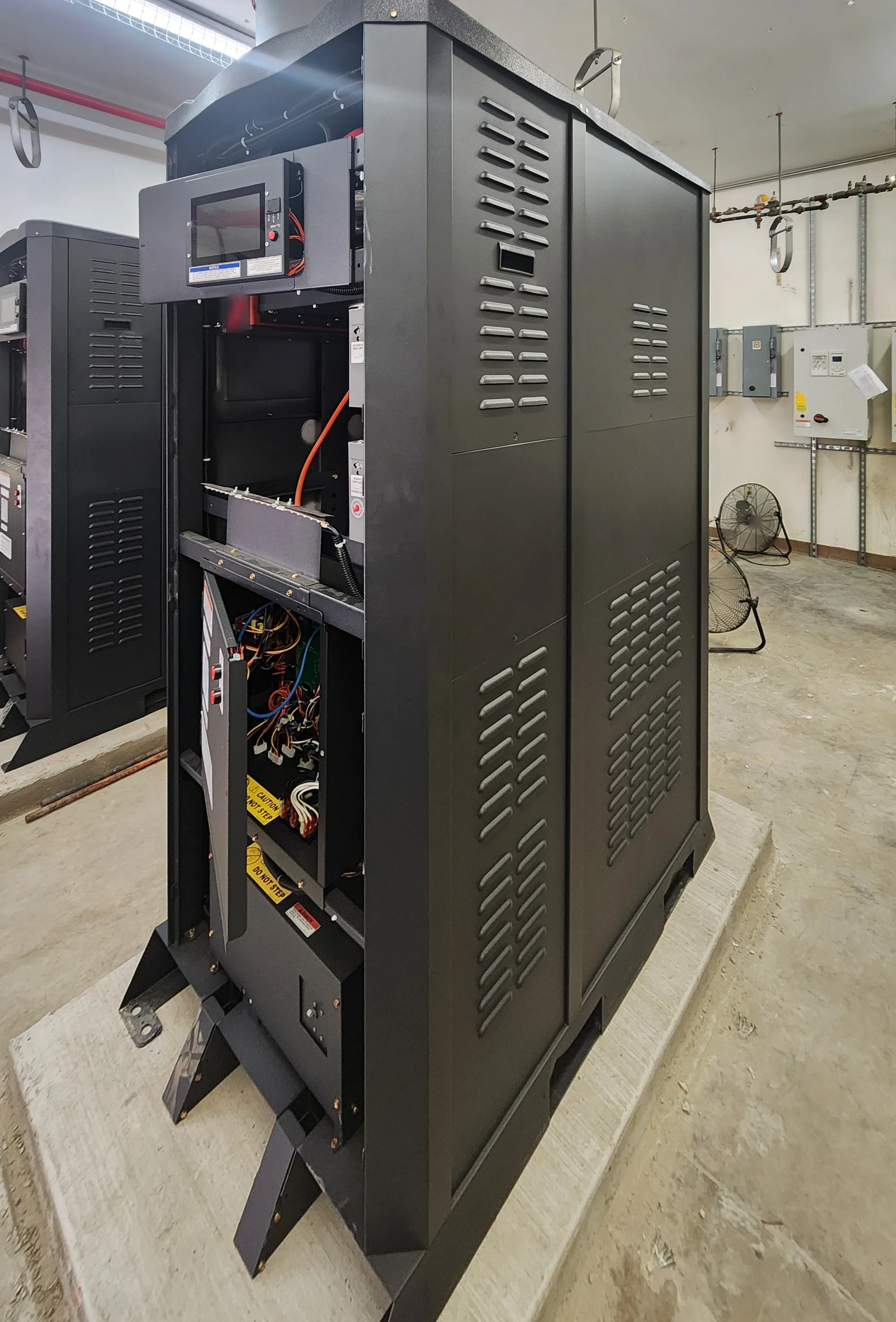 Two tall black boiler cabinets with louvered side panels standing on concrete pads in a boiler room. The near one has its touchscreen control box and a lower control door open, showing wiring; the second stands behind it at the left, and wall mounted drives, disconnects and two floor fans sit along the back wall.