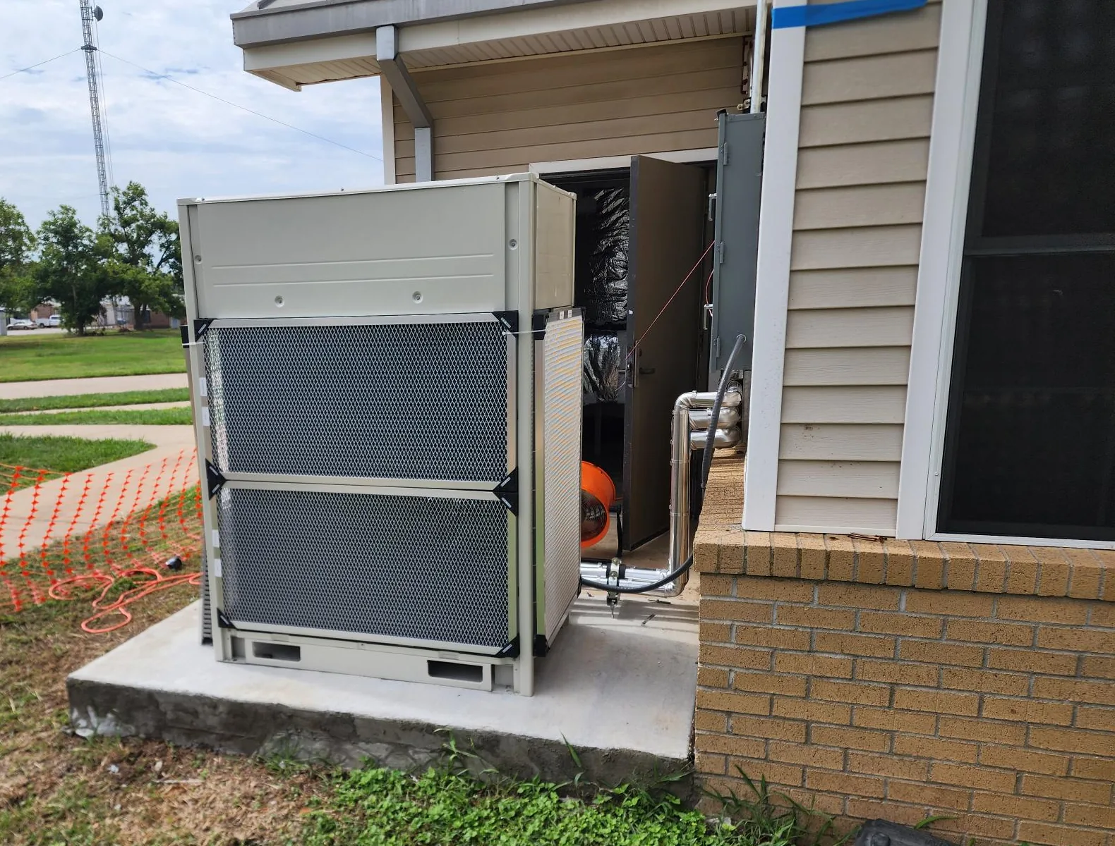 Beige outdoor HVAC unit with mesh coil guards on a fresh concrete pad against a brick and siding building, an open mechanical room door beside it, orange safety fence on the lawn at left and a cloudy sky.